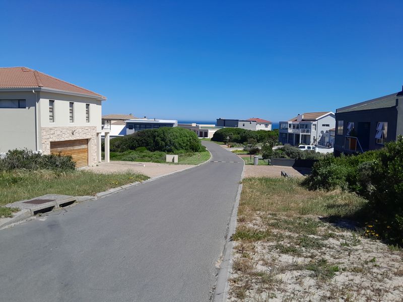A view of a paved residential street in a coastal neighborhood, showing surrounding modern homes and clear blue sky.