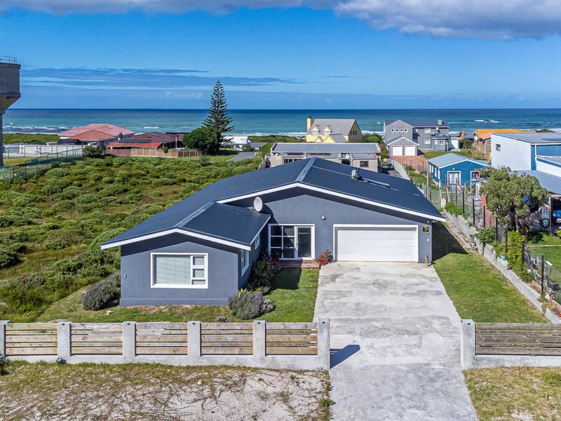 Aerial view of a modern coastal home featuring a dark metal roof, concrete driveway, and secure boundary fencing, situated near the ocean.