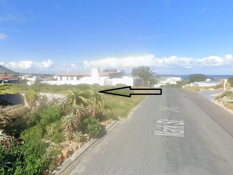 Street view of a vacant residential plot featuring established landscaping and boundary walling in a coastal neighborhood.