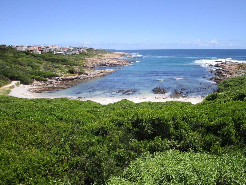 A scenic elevated view of the coastline, featuring a sandy beach, rocky shoreline, and the ocean, with residential properties visible in the distance.