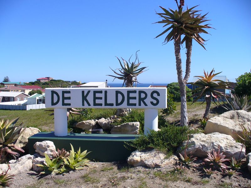 A clear view of the De Kelders area signage set within a landscaped garden with coastal residential properties in the background.