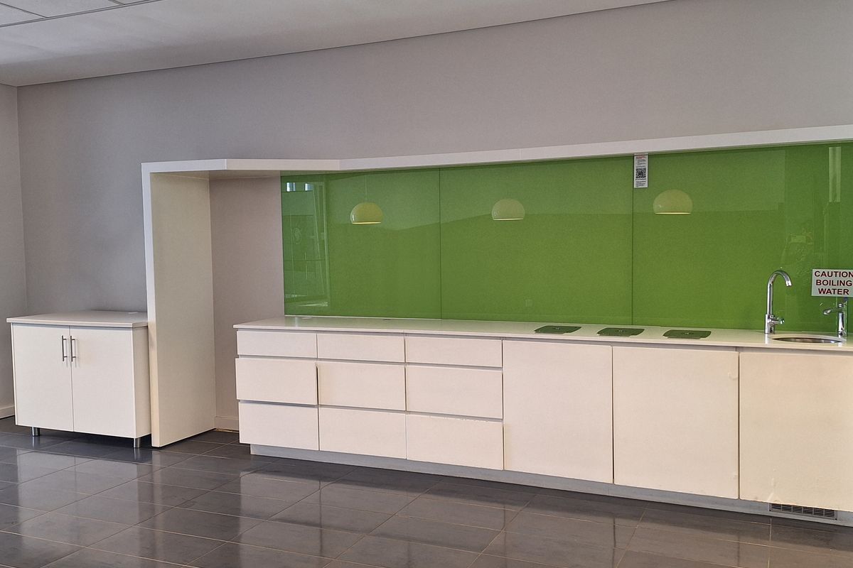 A modern office kitchenette featuring white cabinetry, a vibrant green glass splashback, dark tiled flooring, and a built-in boiling water tap.