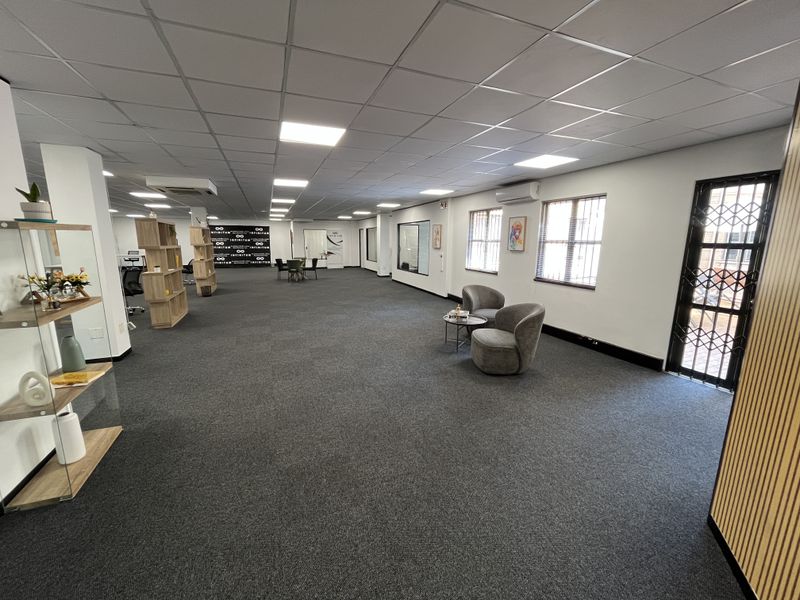 Spacious open-plan office area featuring a suspended ceiling, recessed lighting, carpeted flooring, and secure access points with burglar bars.