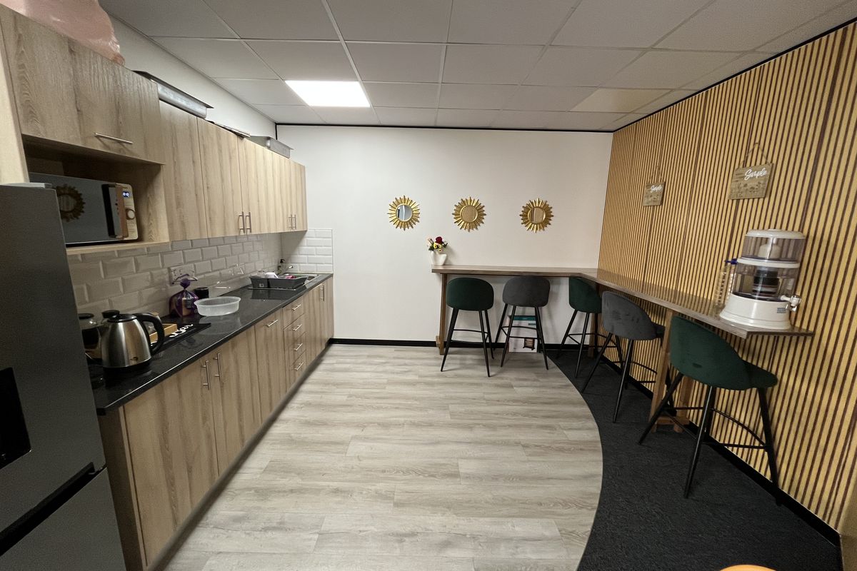 A modern office kitchenette featuring light wood cabinetry, subway tile backsplash, dark countertops, and a dedicated bar-style seating area with slatted wood wall accents.