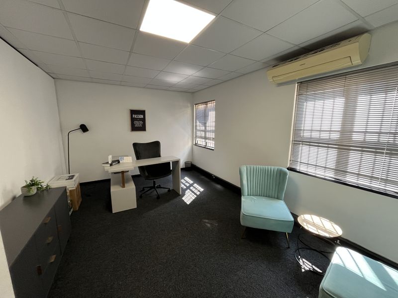 A functional office space featuring a suspended ceiling, recessed lighting, wall-mounted air conditioning, and venetian blinds, finished with dark carpeting.