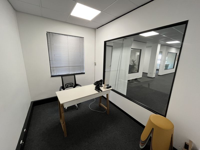 A modern private office space featuring a glass partition wall, carpeted flooring, and recessed ceiling lighting, providing a professional and bright work environment.