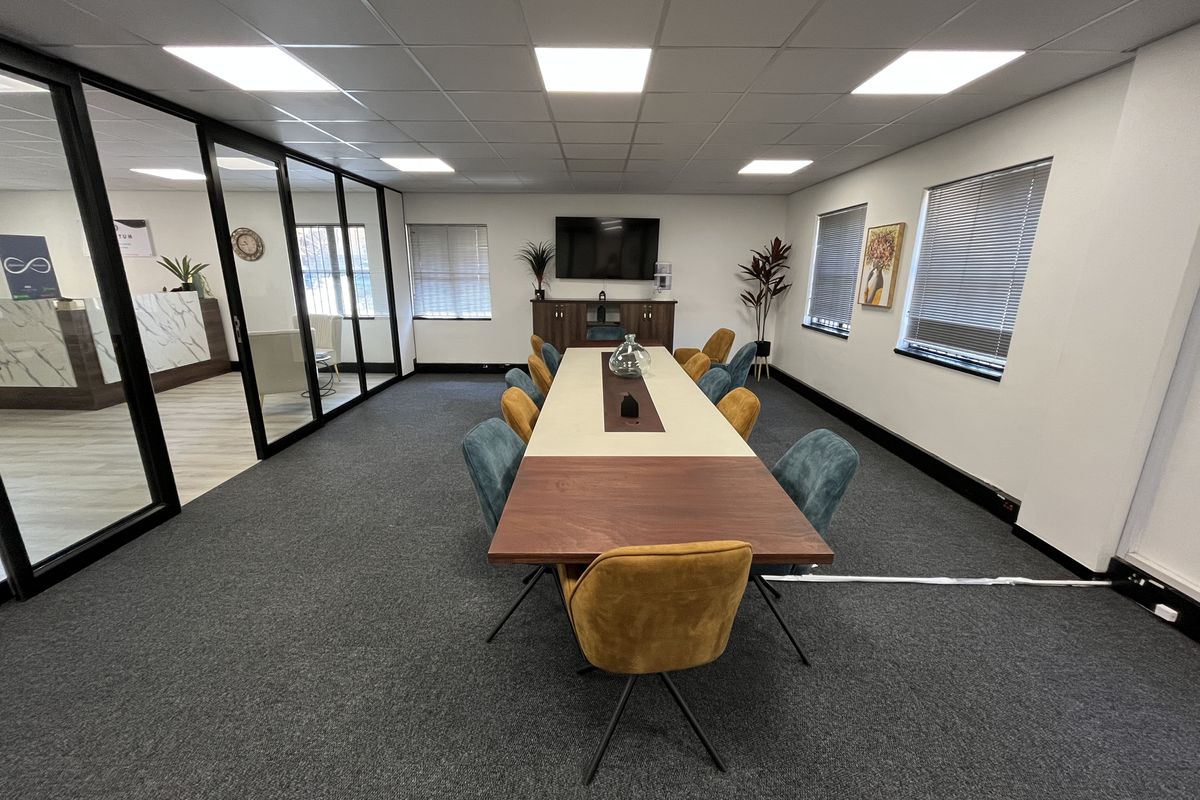 A professional office boardroom featuring a large central table, glass partition walls, suspended ceiling with LED lighting, and carpeted flooring.