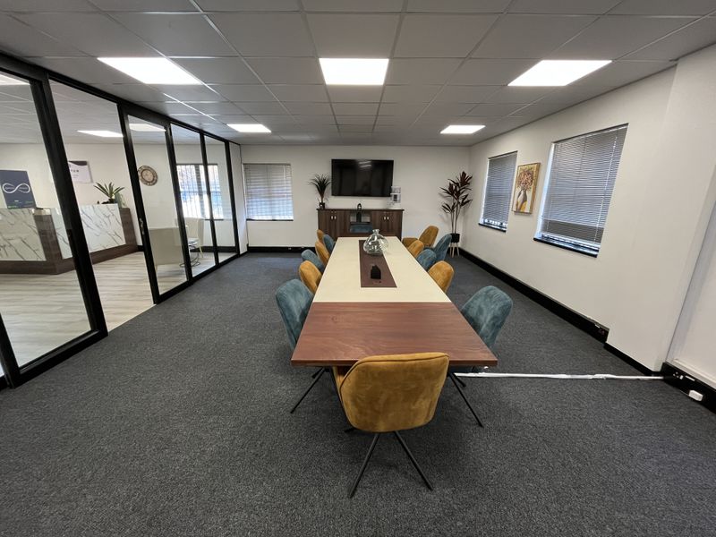 A professional office boardroom featuring a large central table, glass partition walls, suspended ceiling with LED lighting, and carpeted flooring.