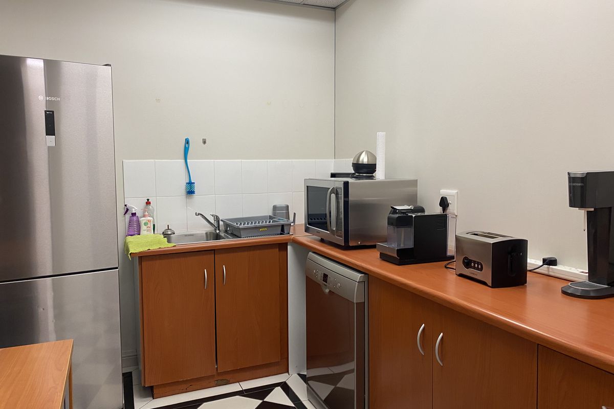A functional office kitchenette featuring wood-finish cabinetry, a stainless steel sink, tiled splashback, and integrated dishwasher space.