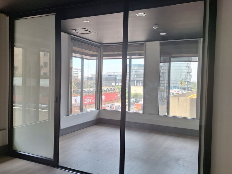 A modern, empty office space featuring large windows, laminate flooring, and a sleek sliding glass partition, offering a bright and professional work environment.