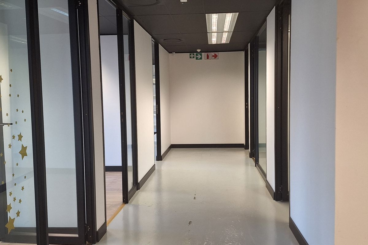 A clean, modern office hallway featuring glass-partitioned rooms, black door frames, suspended ceilings with recessed lighting, and durable epoxy flooring.