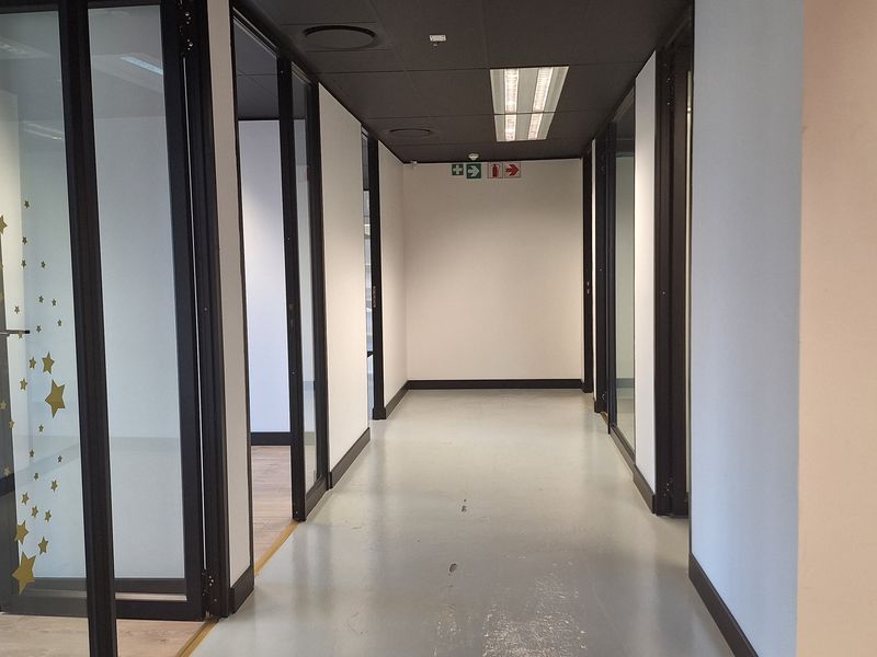 A clean, modern office hallway featuring glass-partitioned rooms, black door frames, suspended ceilings with recessed lighting, and durable epoxy flooring.