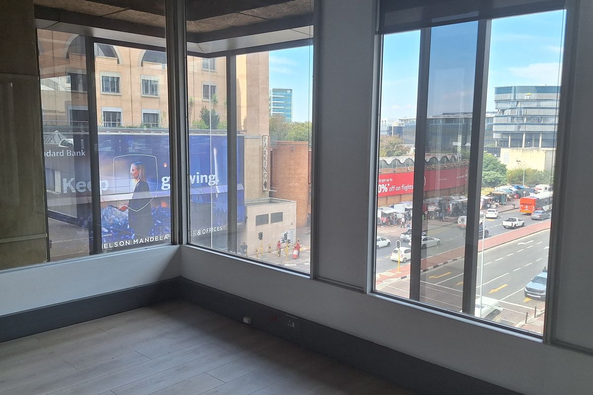 A bright, empty office space featuring large floor-to-ceiling windows with urban views, modern laminate flooring, and clean, neutral finishes.