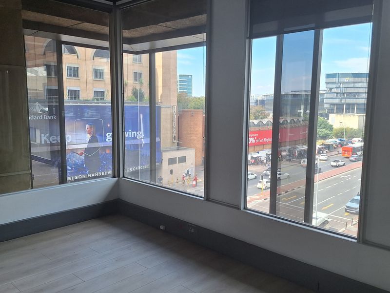 A bright, empty office space featuring large floor-to-ceiling windows with urban views, modern laminate flooring, and clean, neutral finishes.
