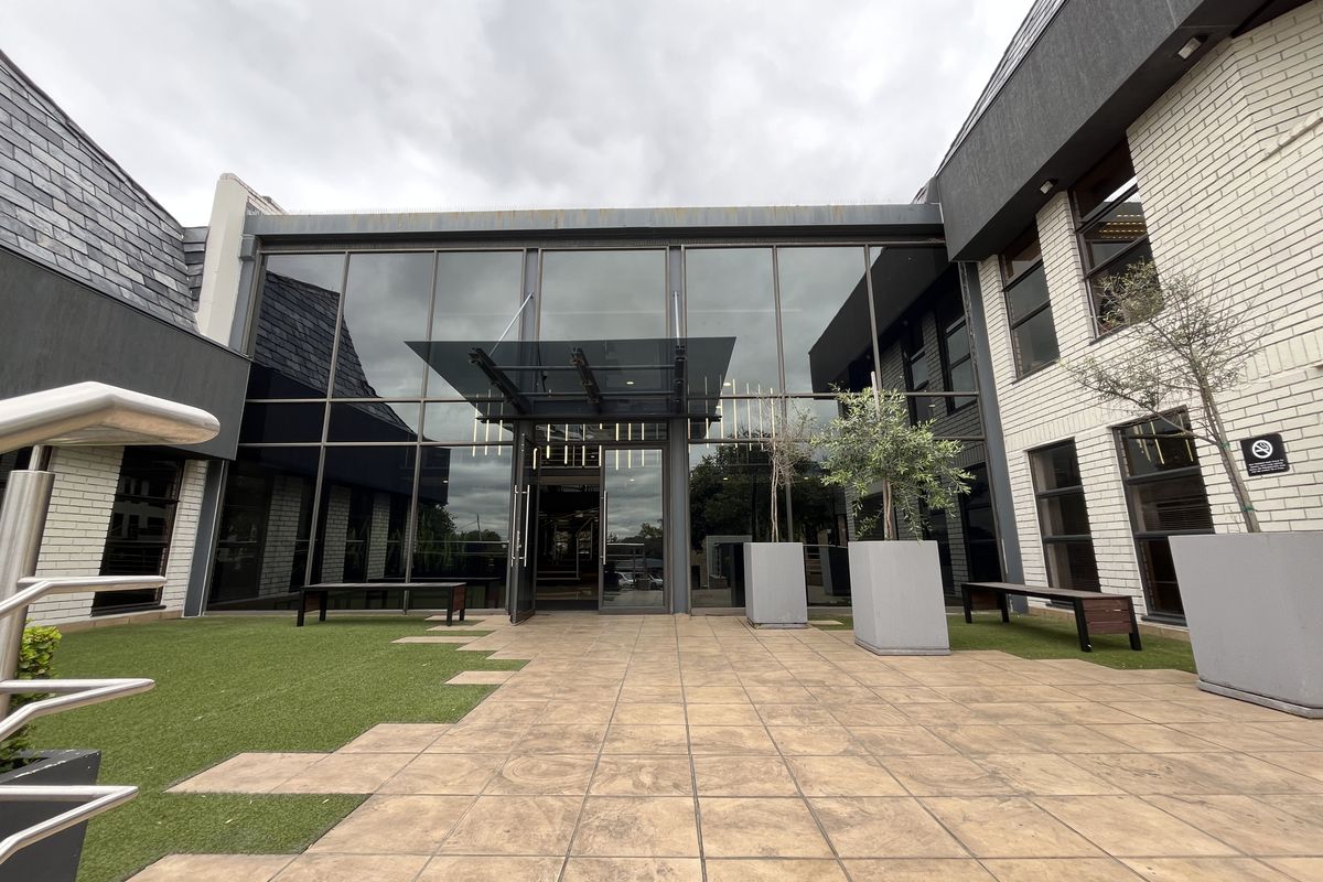 Modern office building entrance featuring a large glass facade, covered entryway, and a paved courtyard area with artificial turf.
