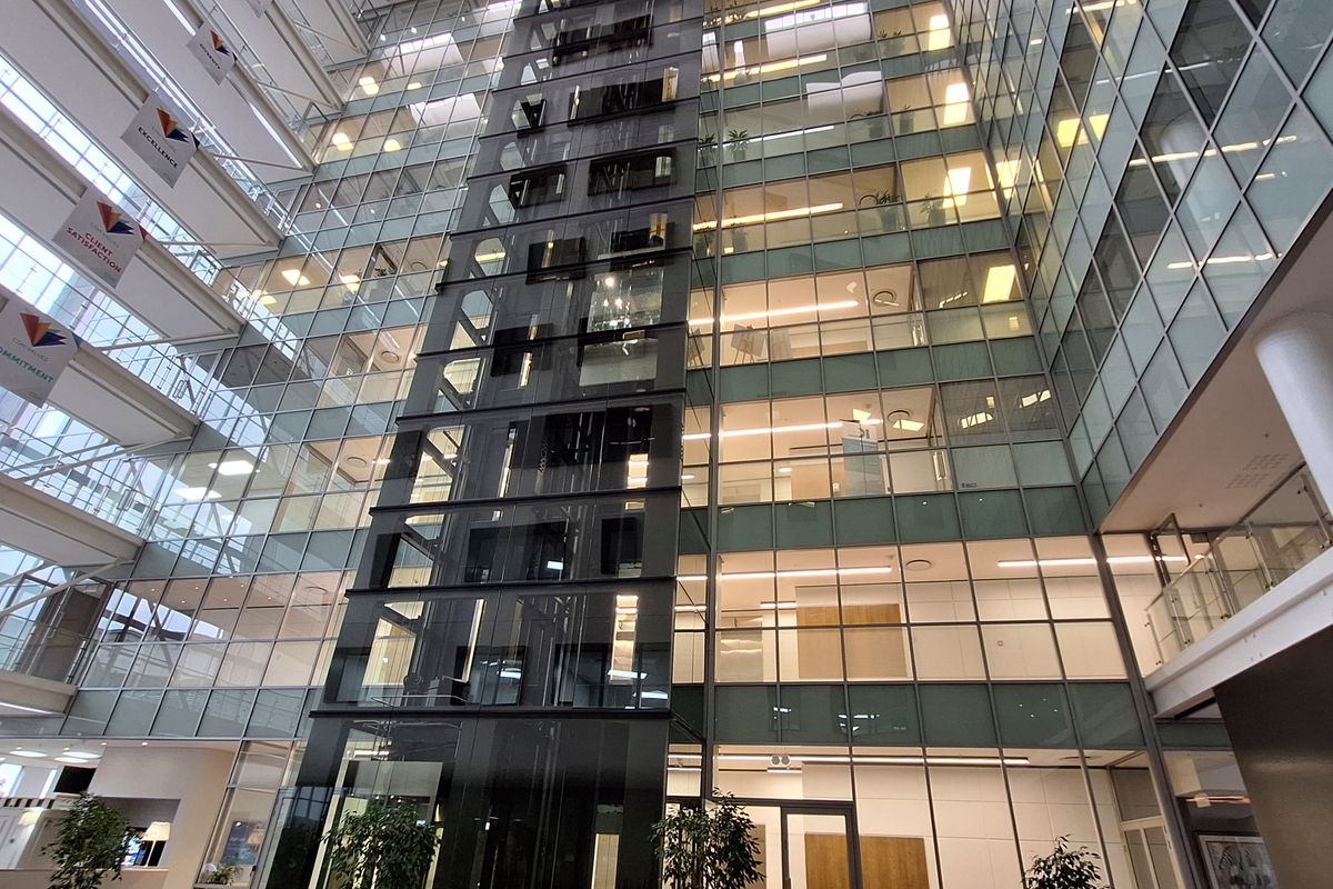 A modern, multi-story office atrium featuring a central glass elevator shaft, expansive glass curtain walls, and natural light from overhead skylights.