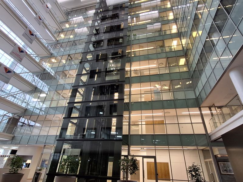 A modern, multi-story office atrium featuring a central glass elevator shaft, expansive glass curtain walls, and natural light from overhead skylights.