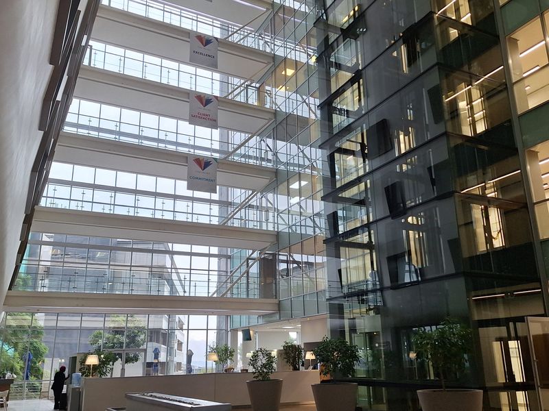 A modern, multi-story office atrium featuring a glass curtain wall, a prominent glass elevator shaft, and a spacious, well-maintained reception area.