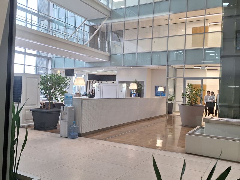 Spacious modern office atrium featuring a high glass curtain wall, polished tiled flooring, and a central reception desk in a professional corporate environment.