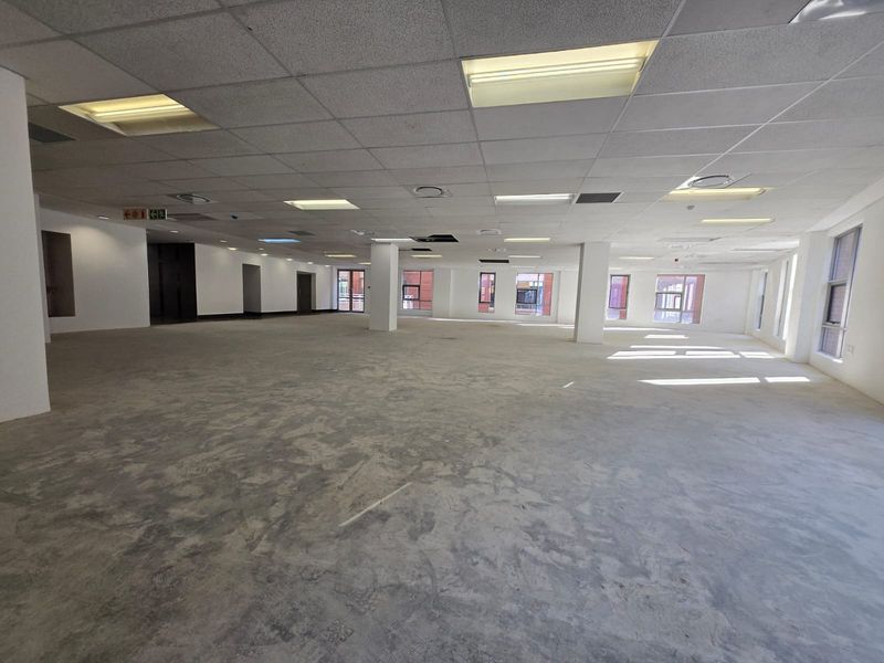 A large, open-plan office space featuring a suspended ceiling, structural columns, and expansive windows, currently in a shell condition awaiting final flooring and fit-out.