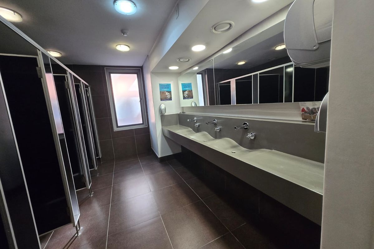 Modern commercial office bathroom featuring a long integrated trough sink, wall-mounted faucets, large vanity mirror, and multiple private toilet cubicles with dark finishes.