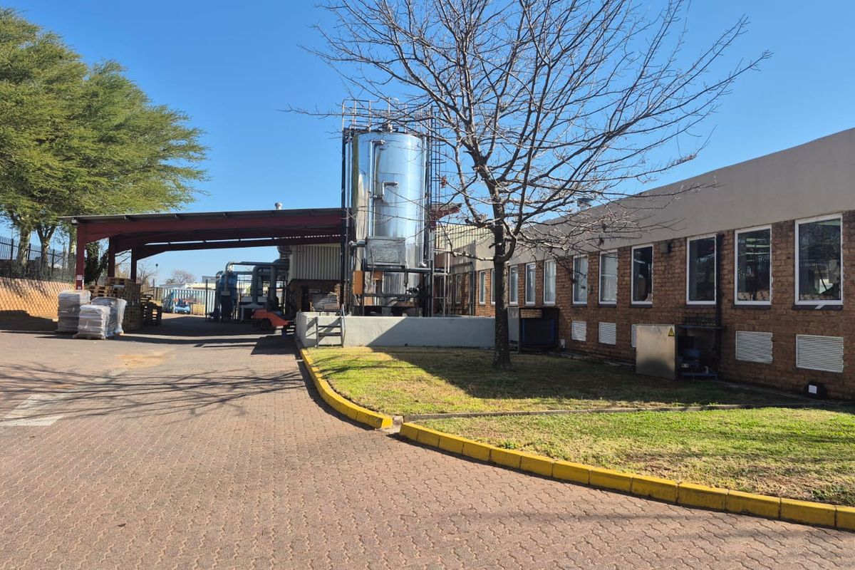 Industrial exterior view featuring a large stainless steel storage tank, covered loading bay, and brick building facade with paved driveway access.