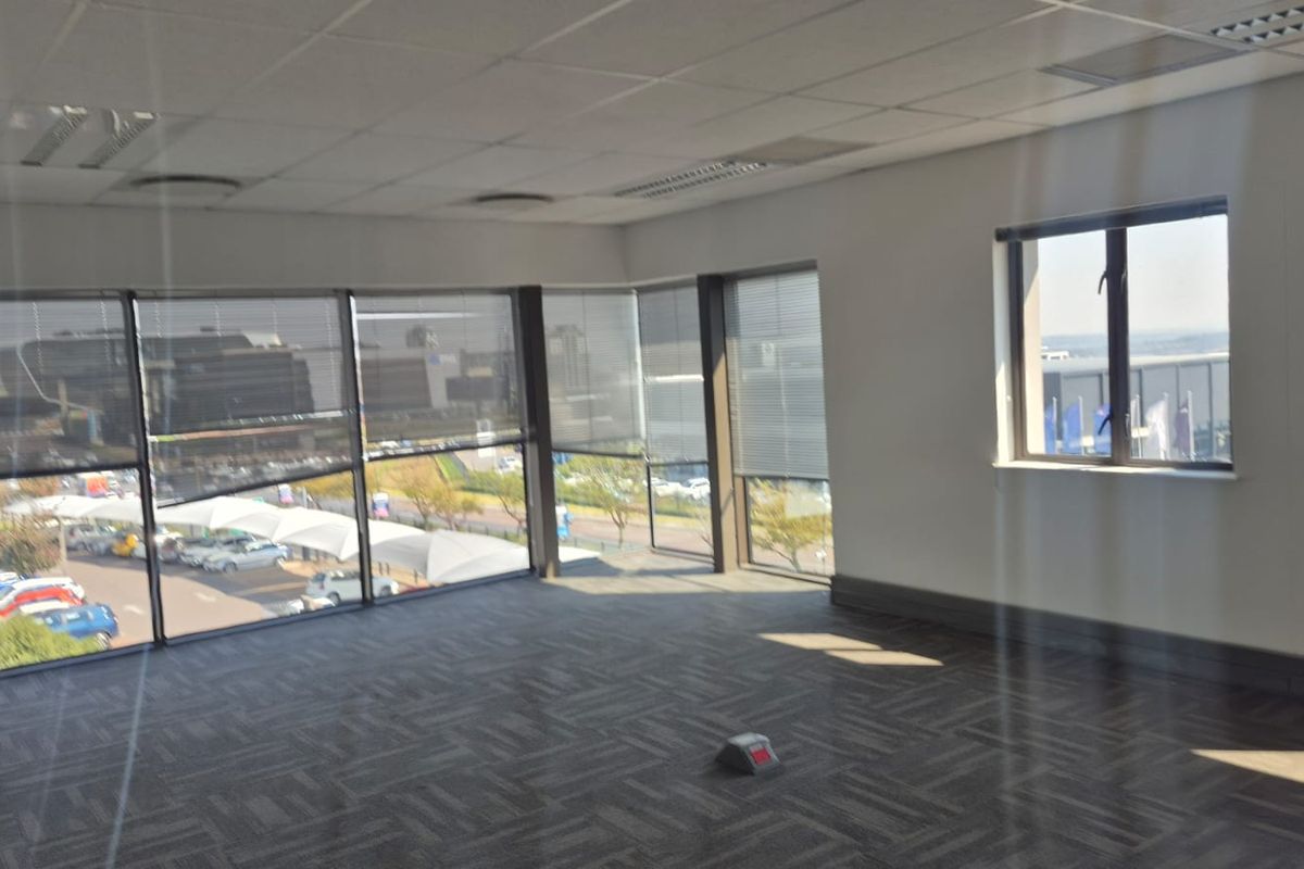 A bright, empty office space featuring large windows with venetian blinds, carpeted flooring, and a suspended ceiling, offering ample natural light and exterior views.