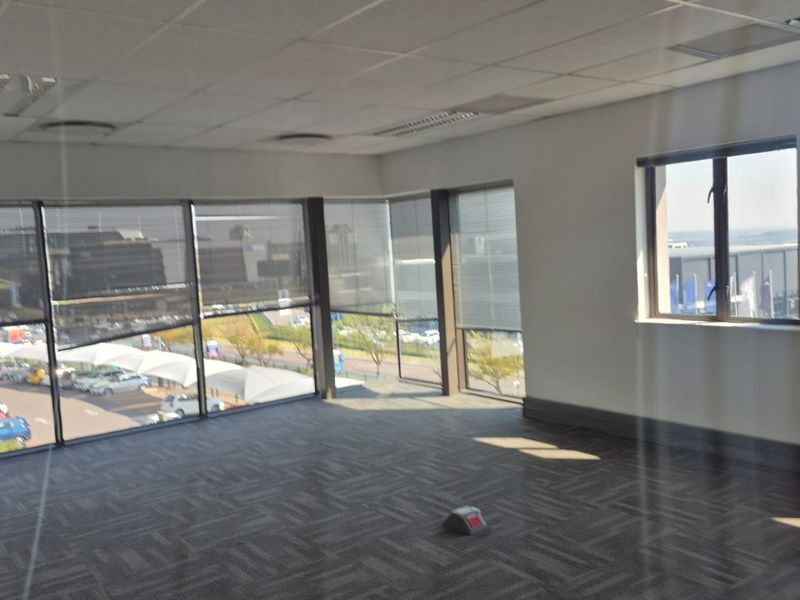 A bright, empty office space featuring large windows with venetian blinds, carpeted flooring, and a suspended ceiling, offering ample natural light and exterior views.
