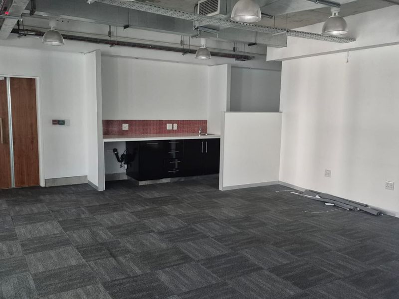 Open plan office space featuring a kitchenette with built-in cabinetry, exposed industrial ceiling, and carpeted flooring.