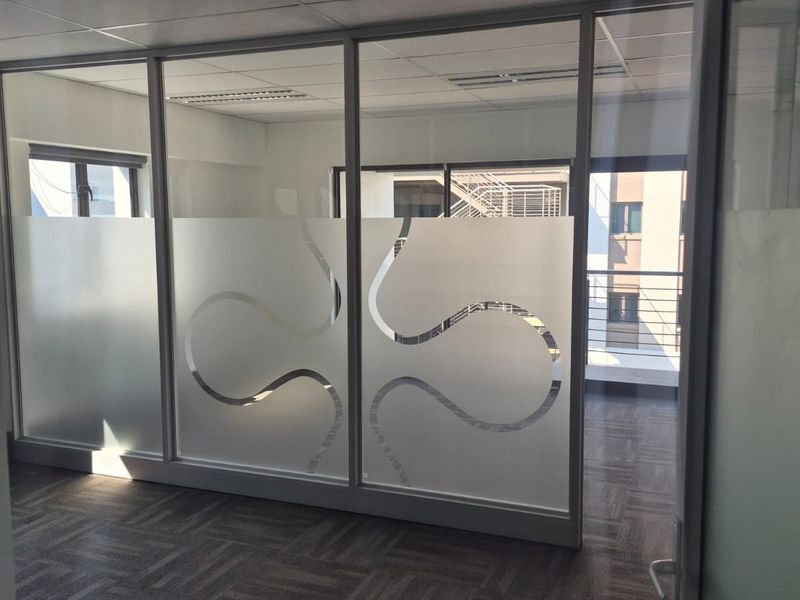 A modern office space featuring glass partitions with decorative frosted film, carpeted flooring, and a suspended ceiling with integrated lighting.