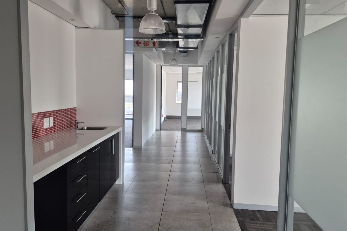 A modern office hallway featuring a built-in kitchenette, tiled flooring, glass partition walls, and exposed industrial-style ceiling ducting.
