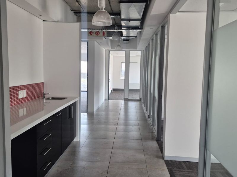 A modern office hallway featuring a built-in kitchenette, tiled flooring, glass partition walls, and exposed industrial-style ceiling ducting.