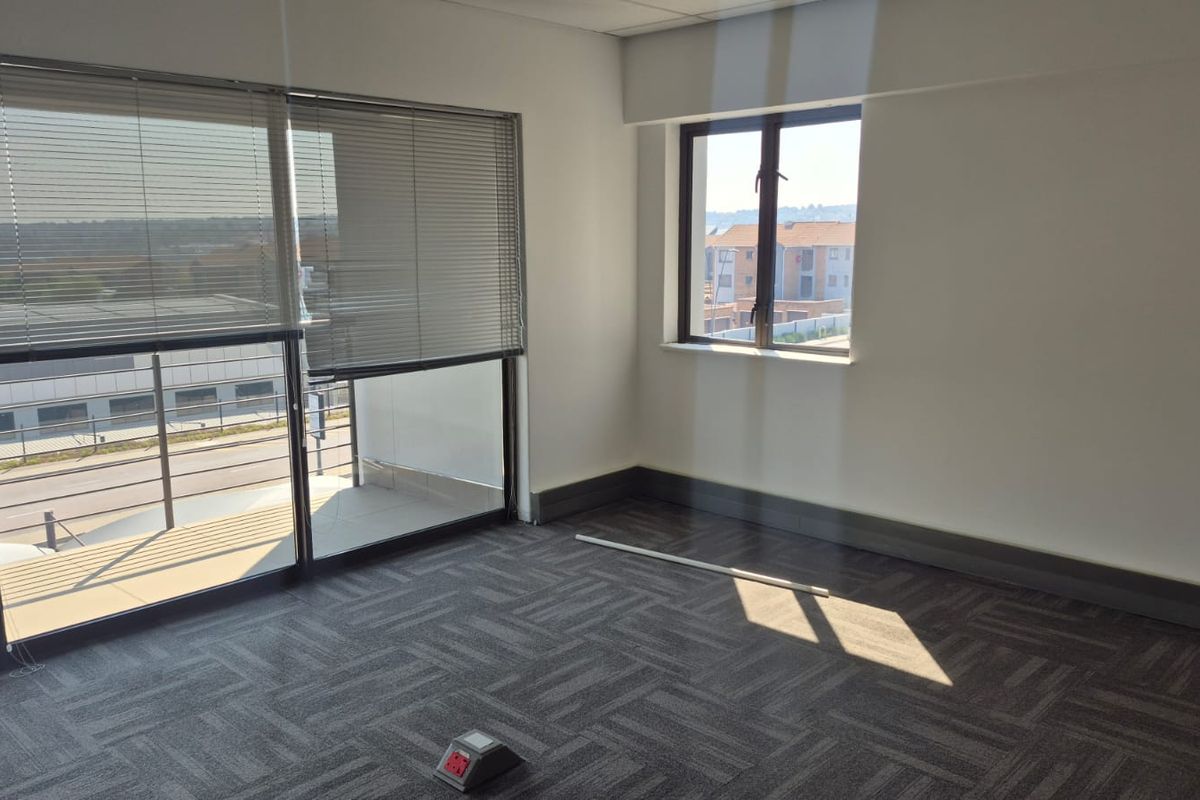 A bright, empty office space featuring carpeted flooring, a floor power outlet, and large windows with venetian blinds providing natural light.