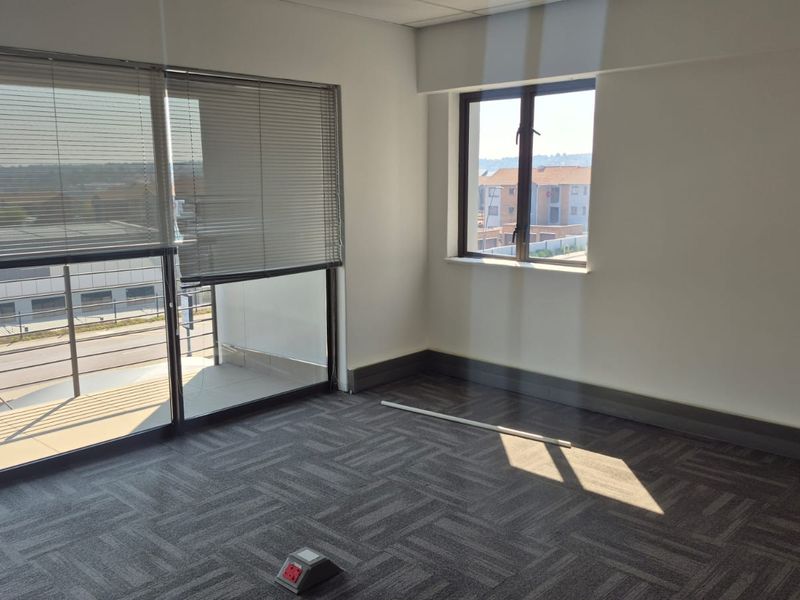A bright, empty office space featuring carpeted flooring, a floor power outlet, and large windows with venetian blinds providing natural light.