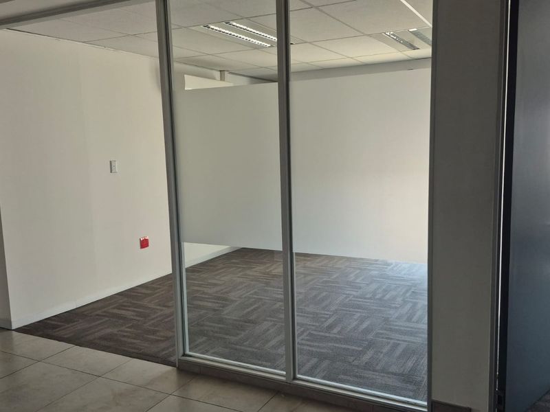 A bright, vacant office space featuring glass partition walls, carpeted flooring, and a suspended ceiling with integrated lighting.