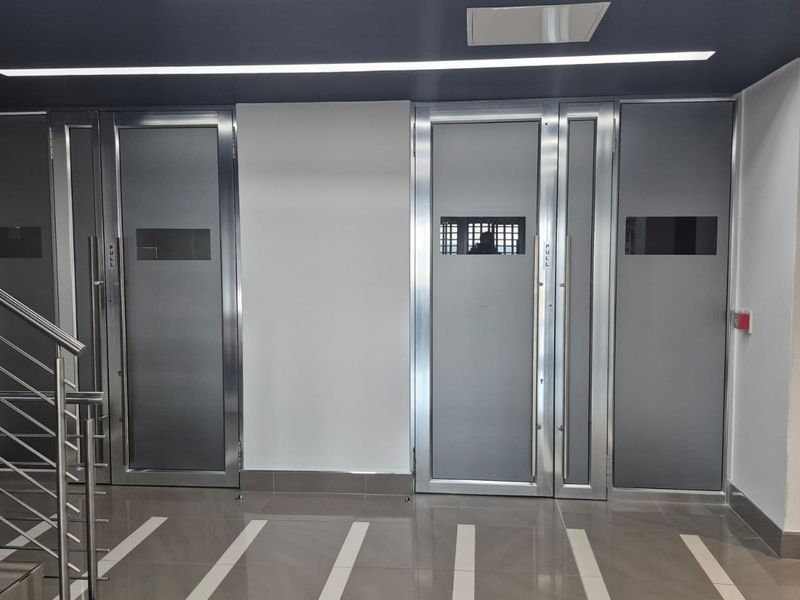 A modern office hallway featuring stainless steel framed glass doors, tiled flooring, and clean architectural lines with recessed lighting.