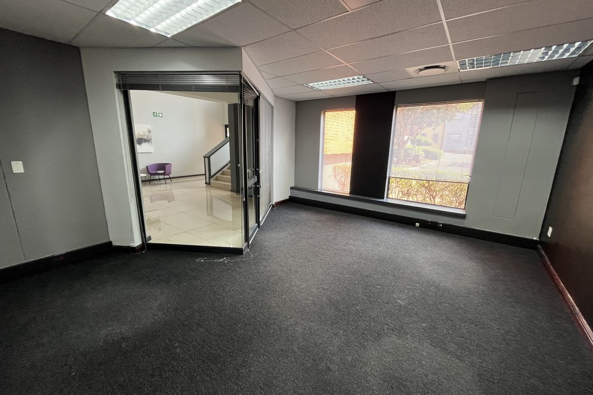 An empty office space featuring dark carpeting, a glass partition wall, and large windows with venetian blinds, providing a professional and functional workspace.