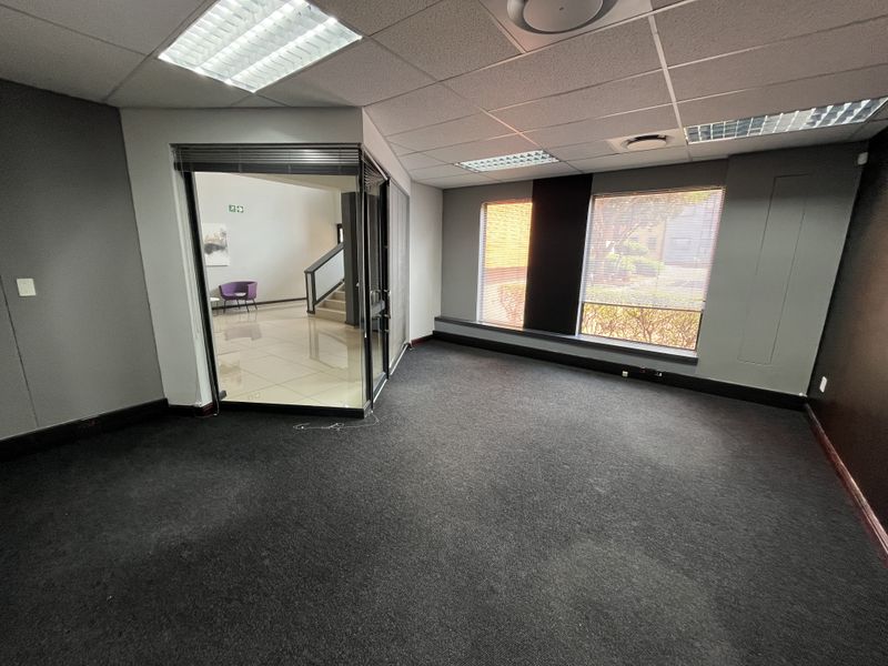 An empty office space featuring dark carpeting, a glass partition wall, and large windows with venetian blinds, providing a professional and functional workspace.