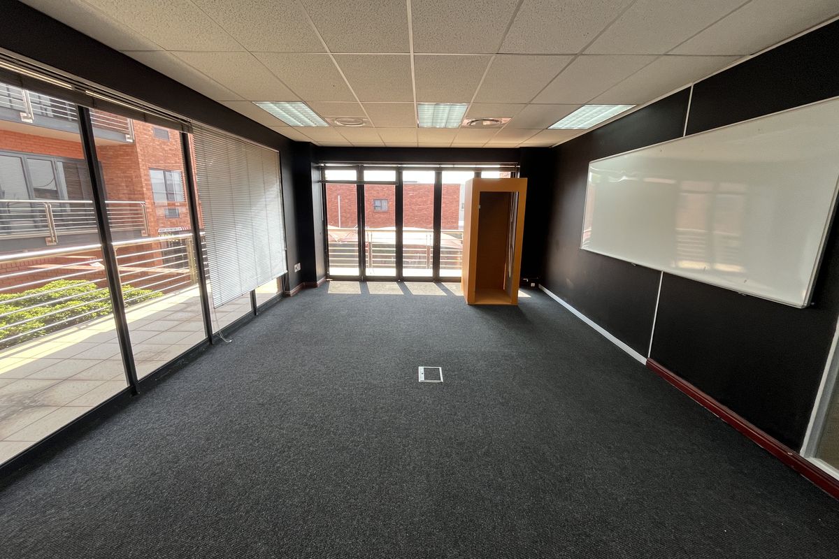 A spacious, empty office featuring dark walls, a suspended ceiling with recessed lighting, floor power access, and large sliding glass doors leading to a balcony.