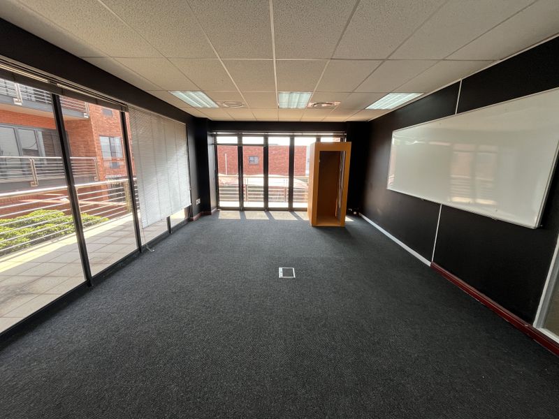 A spacious, empty office featuring dark walls, a suspended ceiling with recessed lighting, floor power access, and large sliding glass doors leading to a balcony.