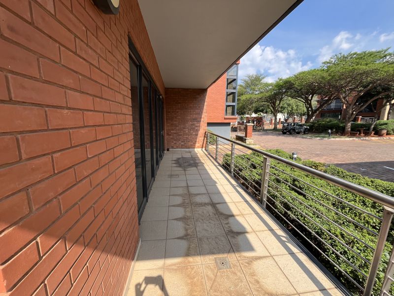 A covered balcony featuring exposed brick walls, stainless steel railings, and tiled flooring, overlooking the office park grounds.