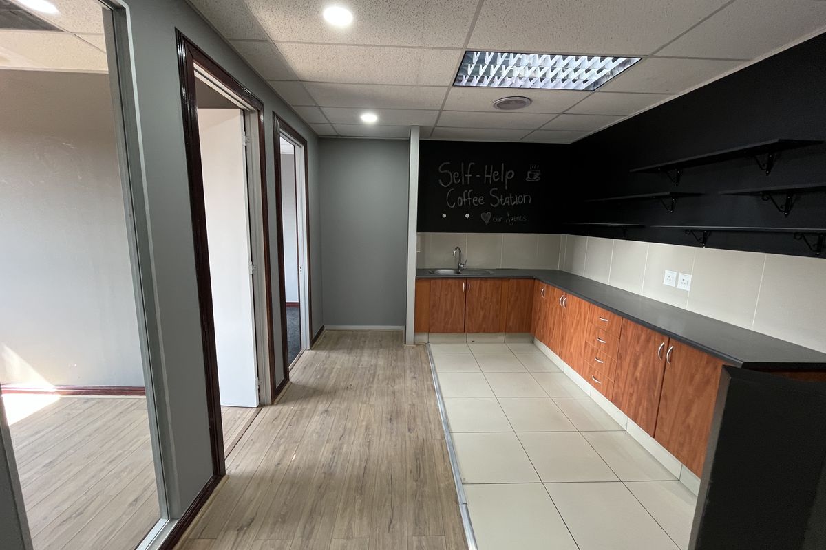 A functional office kitchenette featuring wood-finish cabinetry, a stainless steel sink, laminate countertops, and modern recessed lighting in a clean, open-plan office environment.