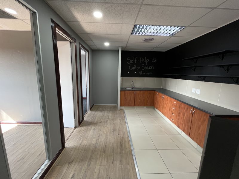A functional office kitchenette featuring wood-finish cabinetry, a stainless steel sink, laminate countertops, and modern recessed lighting in a clean, open-plan office environment.