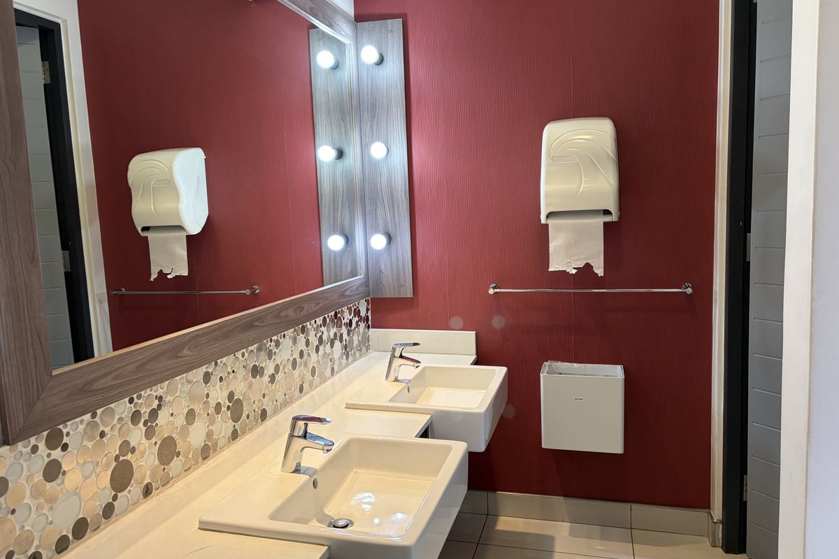 A modern office restroom featuring dual wall-mounted ceramic basins, a large vanity mirror with integrated lighting, and red textured wall finishes.