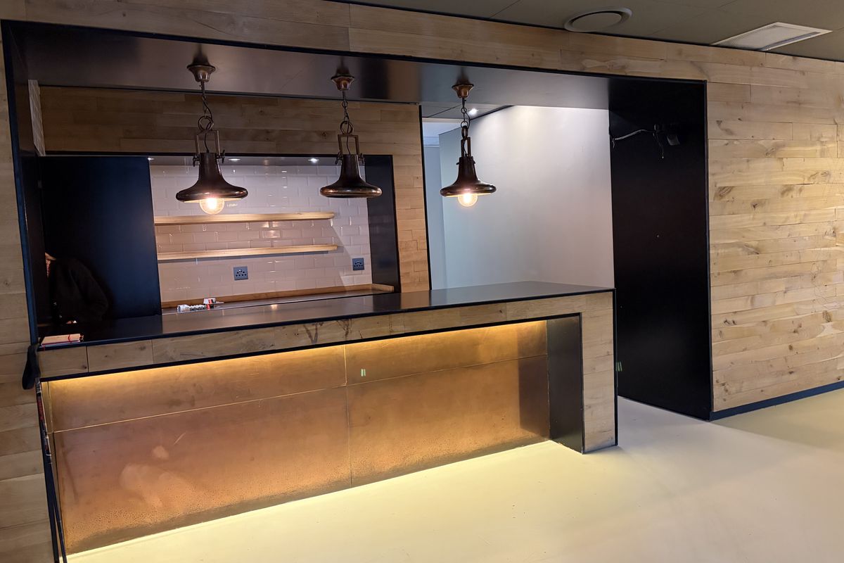 A modern office kitchenette area featuring a sleek copper-fronted counter, industrial-style pendant lighting, and timber-clad wall paneling.