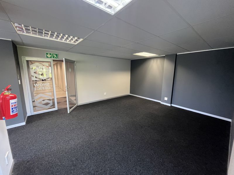 A clean, empty office space featuring dark carpeting, a suspended ceiling with recessed lighting, and a glass-panelled entrance door.