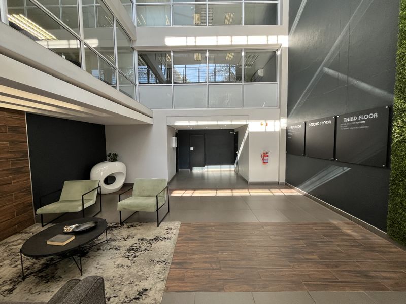 Modern office building entrance lobby featuring high ceilings, large glass windows, tiled flooring, and clear directional signage for multiple floors.