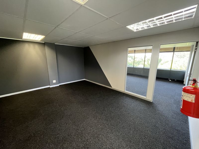 A clean, empty office space featuring dark carpet flooring, a suspended ceiling with recessed lighting, and a glass partition wall.