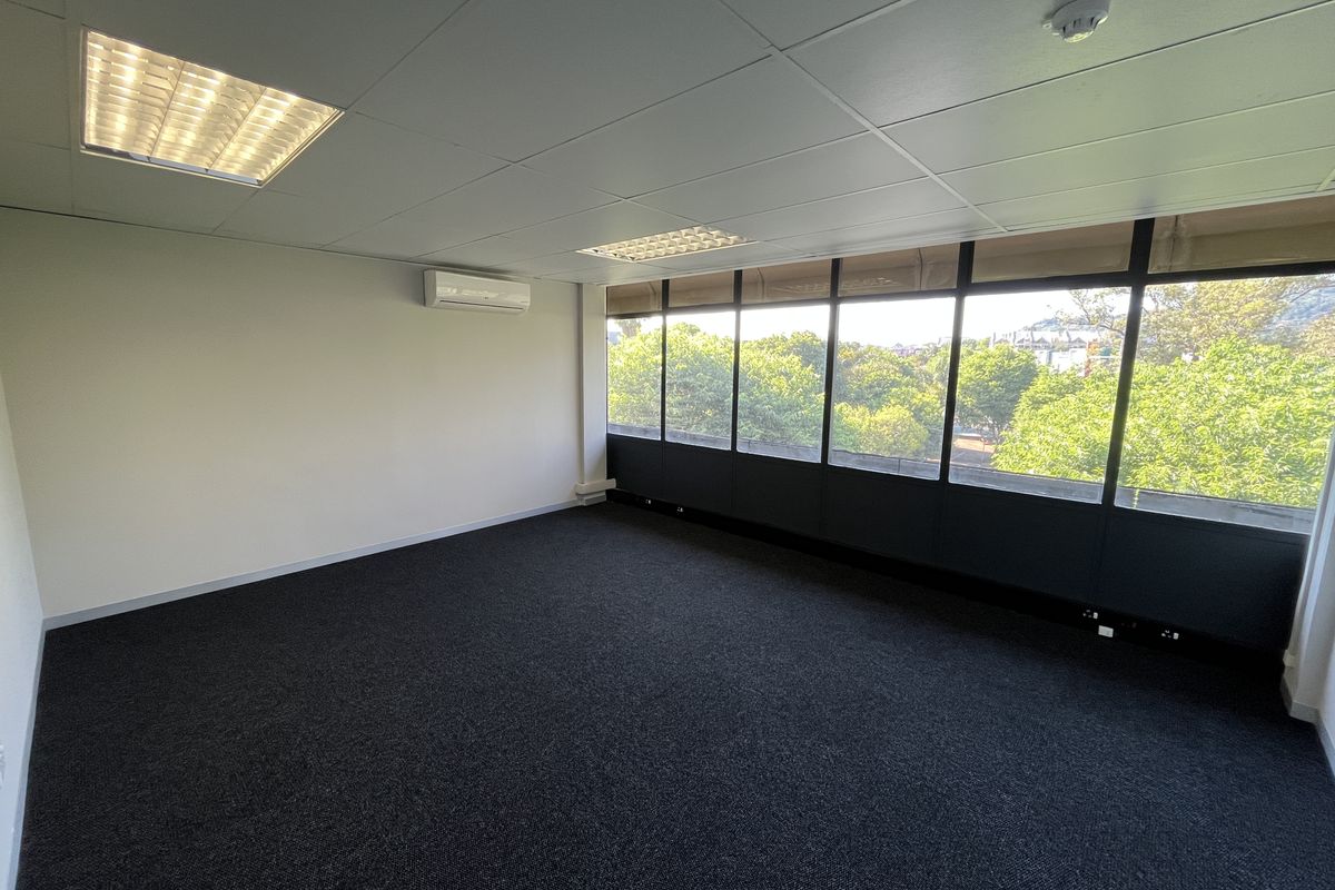 A bright, empty office space featuring carpeted flooring, a suspended ceiling with recessed lighting, a wall-mounted air conditioning unit, and large windows with exterior views.