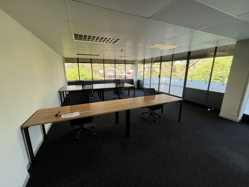 A bright, open-plan office space featuring large windows, suspended ceilings with recessed lighting, dark carpeting, and direct access to an outdoor balcony area.